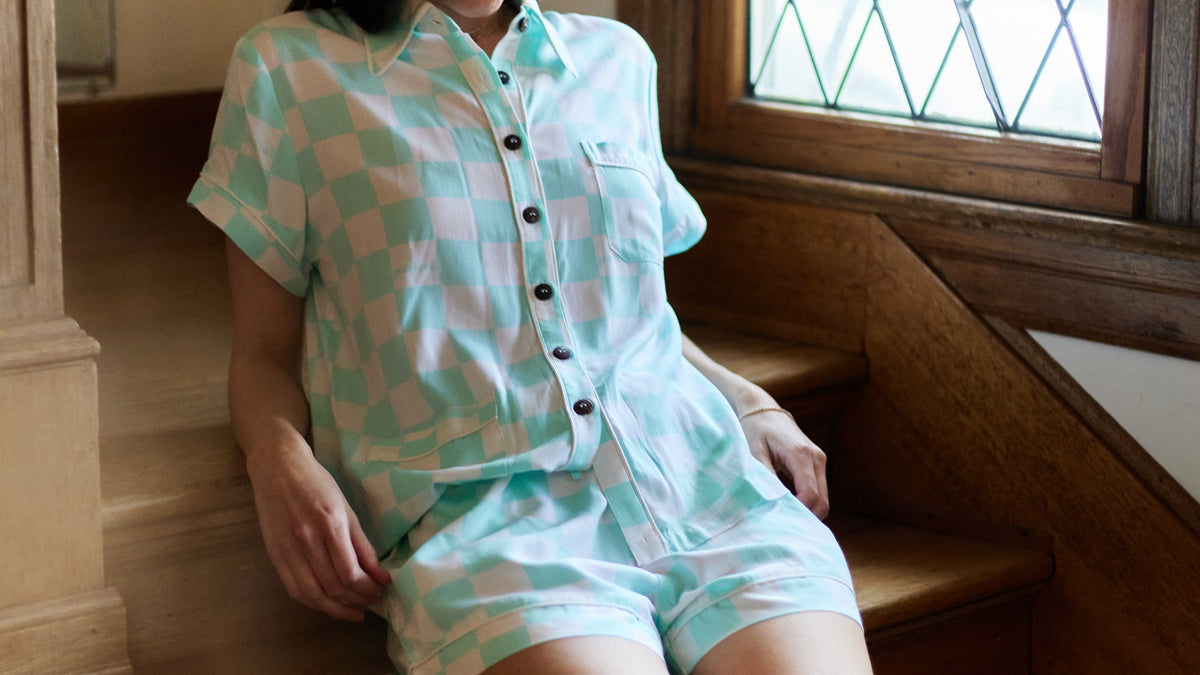 Woman wearing a matching teal and white checkered short-sleeve pajama set sits on a staircase.