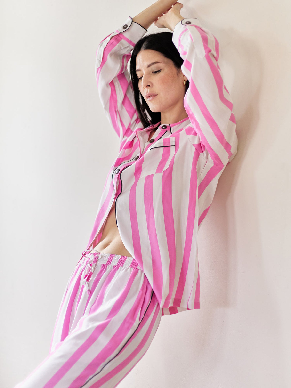 Woman wearing a pink and white striped loungewear set leans against a wall with her arms stretched.