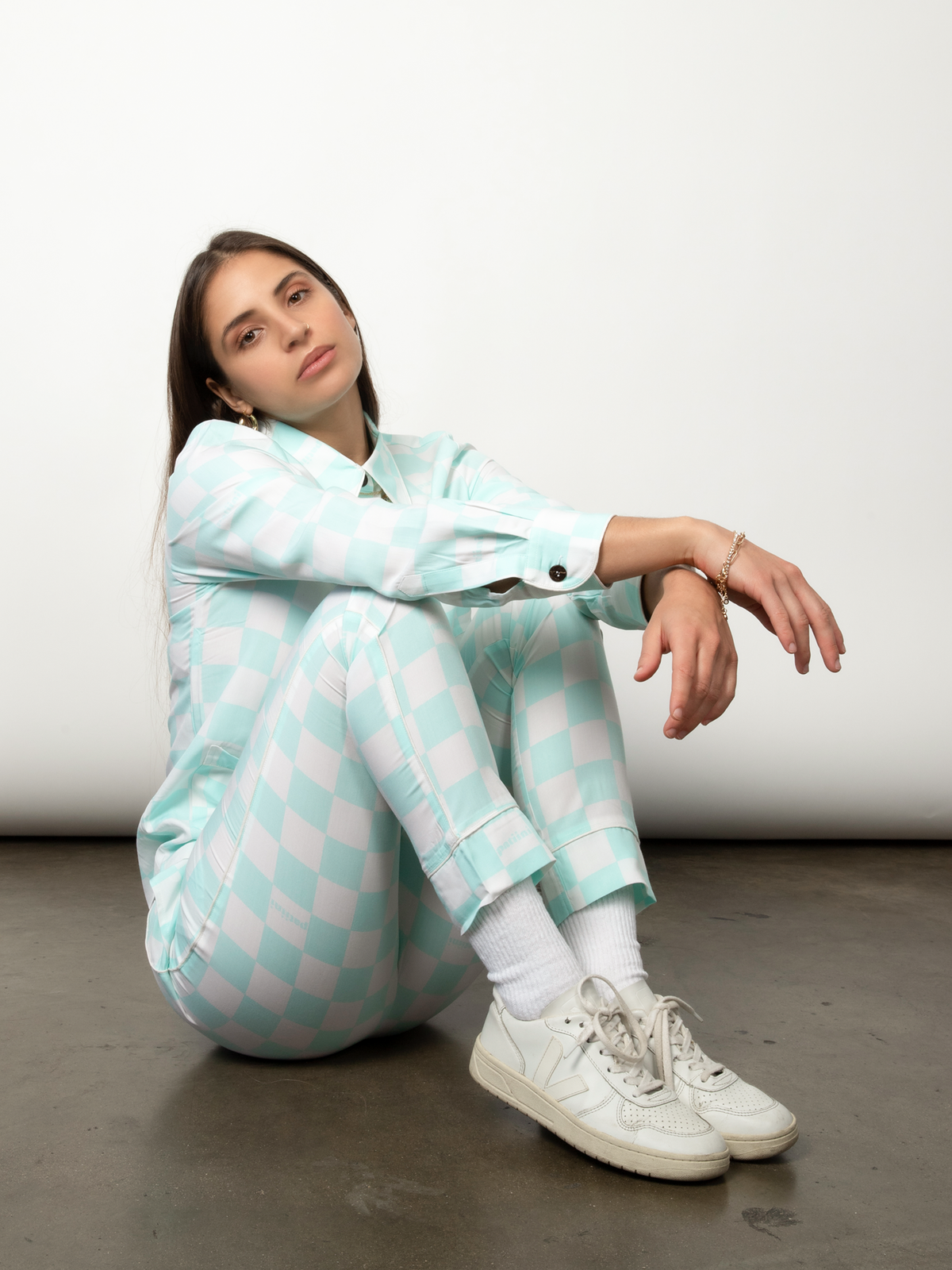 Woman sits with her knees up in a teal and white checkered pajama set.